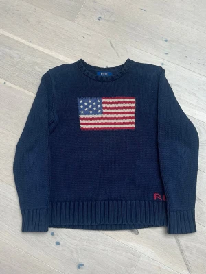 Mörkblå stickad tröja Polo Ralph Lauren - Mörkblå stickad tröja från Polo Ralph Lauren med amerikansk flagga på bröstet och diskret röd RL-brodyr vid nederkanten. Tröjan har rund halsringning och ribbstickade muddar. Tillverkad i bomull, perfekt för en avslappnad och cool stil.