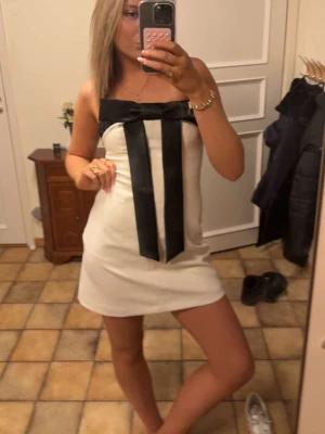 Vit axelbandslös klänning med rosett - Säljer en stilren axelbandslös vit klänning med en stor svart rosett framtill och långa band som detalj. Klänningen är kort och har en rak passform, perfekt för dig som vill sticka ut. Materialet känns mjukt och lätt mot huden.