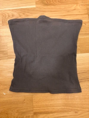 Mörkgrå ribbad tubtopp - Säljer en enkel och stilren mörkgrå tubtopp i ribbat stretchigt tyg. Toppen är axelbandslös och har en rak passform som sitter tajt mot kroppen. Perfekt att matcha med jeans eller kjol för en clean look.