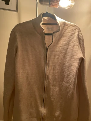 Beige ziptröja från These Glory Days - Snygg beige ziptröja från These Glory Days med hög krage och diskret rutstruktur i materialet. Tröjan har dragkedja framtill och ribbade detaljer vid krage och ärmslut. Perfekt för lager på lager och enkel att matcha med olika outfits.