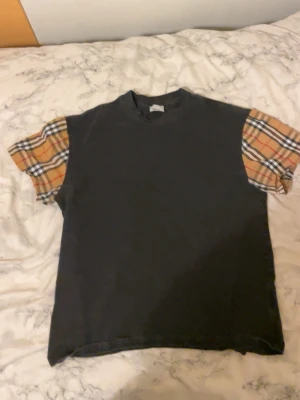 Svart t-shirt med rutiga ärmar - Unik svart t-shirt med klassiska beige-rutiga kortärmar från Burberry. Rak passform och rund halsringning. Perfekt för dig som vill sticka ut nu till våren/sommaren