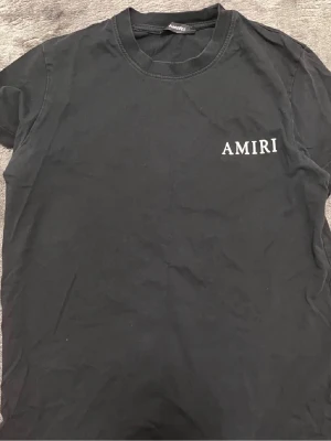 Amiri t shirt  - Svart t-shirt från AMIRI med klassisk passform. Framsidan har en diskret AMIRI-logga på bröstet och baksidan pryds av ett stort tryck med en ängel och AMIRI-text. Perfekt för dig som gillar streetwear med exklusiv känsla.