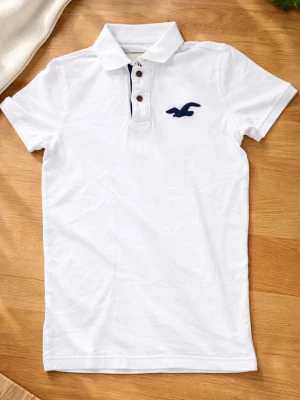 Vit pikétröja från Hollister - Klassisk vit pikétröja från Hollister med korta ärmar och blå logga broderad på bröstet. Tröjan har krage och två knappar framtill. Perfekt till jeans eller shorts för en clean och enkel stil.