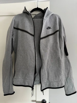 Nike tech fleece  - Snygg grå Nike teech fleece från Nike med svart logga på bröstet och svarta detaljer längs dragkedjan och ärmarna. Hoodien har hel dragkedja, huva och ribbade muddar. Perfekt för en sportig och avslappnad stil. Lite sönder i byxorna men inget som syns när man bär den! 