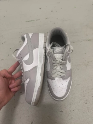 Nike Dunk Low grå och vita sneakers - Nike Dunk Low sneakers i grått och vitt med klassisk Swoosh på sidan. Skorna har perforerad tåbox, vita skosnören och en vit sula med grå detaljer. Tillverkade i läder och textil för en clean och stilren look.
