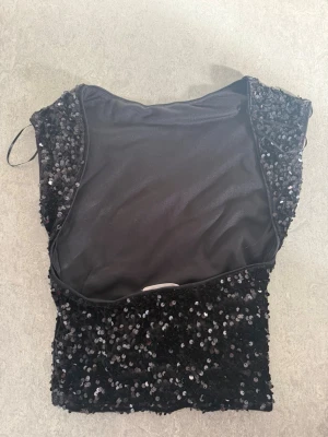 Svart paljettopp croppad - Säljer en svart croppad topp täckt med glittriga paljetter. Toppen har en tight passform och är ärmlös, perfekt för att sticka ut på festen. Materialet känns mjukt mot huden och ger en snygg siluett. 