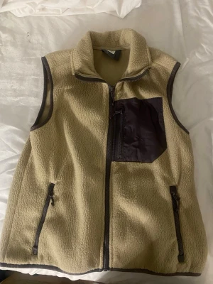 Beige fleeceväst med bröstficka - Beige fleeceväst med dragkedja framtill och kontrasterande mörkbrun bröstficka i nylon. Två sidofickor med dragkedja och hög krage för extra komfort. Perfekt för lager-på-lager och en avslappnad stil.