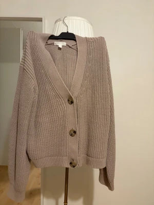 Beige stickad kofta med stora knappar - Säljer en beige stickad kofta med djup V-ringning och tre stora bruna knappar framtill. Koftan har ribbade muddar och en avslappnad passform, perfekt för lager på lager. Passar dig som gillar en enkel men snygg stil.