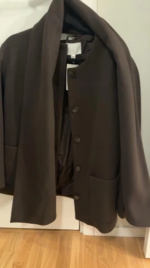 Mörkbrun kappa med stora fickor - Stilren mörkbrun kappa med rund halsringning,  stora knappar framtill samt en scarf. Kappan har två rymliga fickor och en rak passform. Tillverkad i ett mjukt tyg som ger ett elegant intryck. Aldrig använd med prislapp kvar! 