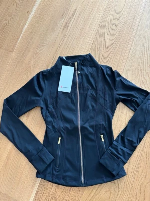 Svart träningsjacka från Lululemon - Snygg svart träningsjacka från Lululemon med figurnära passform, hel dragkedja framtill och två fickor med guldfärgade dragkedjor. Tillverkad i ett stretchigt och funktionellt material som passar perfekt till träning eller som ett extra lager. STORLEK 4
