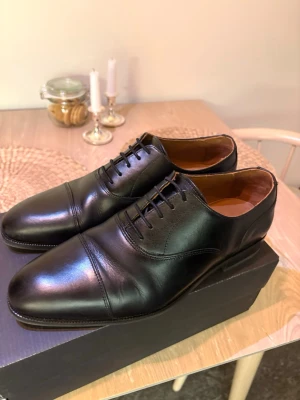 Svarta klassiska Oxfords i läder - These Glory Days oxfords i svart färg, storlek 44. Klassisk snörmodell med tåhätta. Ovandel i slätt material med lätt glans. Passar som dressade skor till kostym eller annan mer formell klädsel.  Köpta på Volt för 3 månader sen - använda 2ggr.  Nypris 1599kr - låda medföljer ej.  Frakt möjlig, annars hämtas de i Hägersten