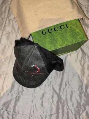 Gucci keps med orm och GG-mönster - Snygg svart keps från Gucci med klassiskt GG-monogram och en rödvit orm broderad framtill. Kepsen har mesh på sidorna och justerbar rem bak. Materialet är en mix av bomull och polyester. Perfekt för dig som vill sticka ut med en lyxig accessoar. 