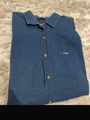 Blå skjorta från U.S. Polo Assn - Snygg blå skjorta från U.S. Polo Assn med diskret mönster och klassisk krage. Skjortan har knappar framtill och en liten logga på bröstet. Modellen är slim fit och passar perfekt till jeans eller chinos.