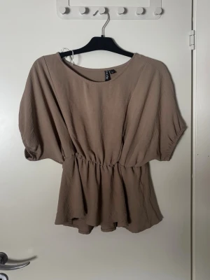 Beige topp med resår i midjan från Pieces - Säljer en beige topp från Pieces med lös passform, resår i midjan och korta, vida ärmar. Toppen har rund halsringning och ett mjukt, lätt material som ger ett snyggt fall. Perfekt att styla med jeans eller kjol.