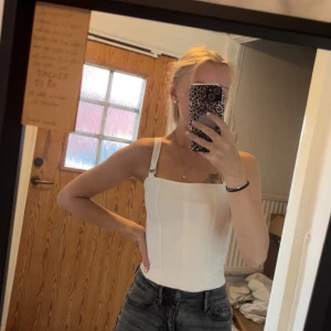 Korsettinspirerad bodysuit - Säljer en vit och en svart korsettinspirerad bodysuit från H&M med breda axelband och markerade sömmar framtill. Bodyn har fyrkantig ringning och knäppning i grenen. Perfekt att styla med jeans eller kjol för en clean look. Säljer två likadana, en svart & en vit.