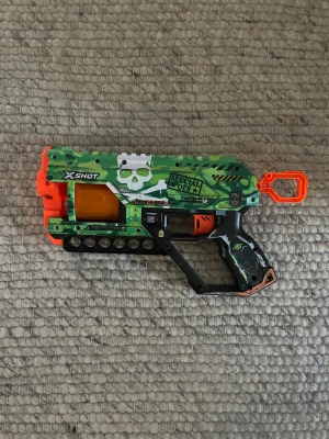 X-Shot leksakspistol Special Ops - Cool leksakspistol från X-Shot med kamouflagemönster i grönt, svart och orange. Pistolen har en stor vit dödskalle på sidan, texten 'Special Ops' och plats för skumdartpilar. Greppet är svart med detaljer i grönt och orange. Har knappt använt, jag har inga skott till den. 