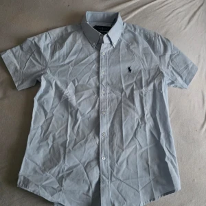 Ljusblå kortärmad skjorta Polo Ralph Lauren - Klassisk ljusblå skjorta från Polo Ralph Lauren med korta ärmar och button-down krage. Skjortan har knappar framtill och den ikoniska loggan broderad på bröstet. Perfekt till jeans eller chinos för en clean och stilren look.