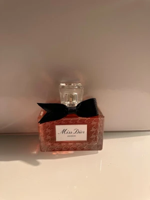 Miss Dior Essence parfym - Miss Dior Essence parfym i en rektangulär glasflaska med ett elegant mönster och ljusrosa doftvätska. Flaskan har en genomskinlig fyrkantig kork och en svart rosett runt halsen, vilket ger en lyxig och feminin känsla. 80 ml. Ordinarie pris 2369kr.