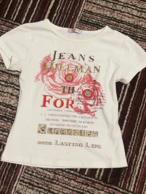 Vit t-shirt med tryck  - Säljer en vit t-shirt  med färgglatt tryck framtill. T-shirten har en stor röd ros, guldfärgade bokstäver och flera dekorativa detaljer. Rund halsringning och korta ärmar. 