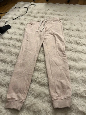 Ljusrosa mjukisbyxor med dragsko - Mysiga ljusrosa mjukisbyxor med resår i midjan och dragsko för perfekt passform. Byxorna har ribbade muddar vid bensluten och är tillverkade i ett mjukt, bomullsliknande material. Perfekta för chill dagar hemma eller när du vill vara extra bekväm.