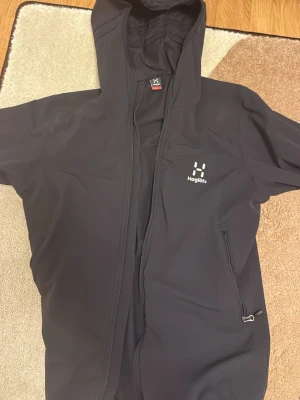 Svart vindjacka från Haglöfs med huva - Snygg svart vindjacka från Haglöfs med dragkedja framtill, justerbar huva och två sidofickor med dragkedja. Jackan har minimalistisk design och diskret Haglöfs-logga på bröstet. Perfekt för blåsiga dagar och outdoor-aktiviteter.