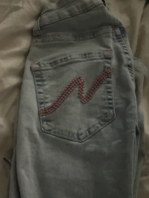 Ljusblå slitna jeans med broderad ficka - Säljer ett par ljusblå jeans med raka ben och slitningar vid knäna. På bakfickan finns ett rosa broderat mönster som ger en unik touch. Jeansen har klassisk femficksmodell och är tillverkade i denim. Perfekta för en avslappnad och trendig look. Från nelly!!