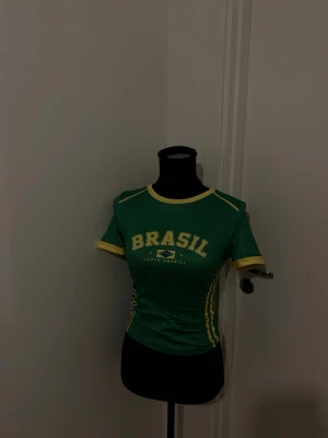 Grön Brasil topp med gula detaljer - Sportig grön topp med texten 'BRASIL' och brasilianska flaggan framtill. T-shirten har gula detaljer vid halsringning, ärmslut och längs sidorna. Kortärmad och figurnära passform, perfekt för dig som gillar färgstarka plagg med sportig vibe.