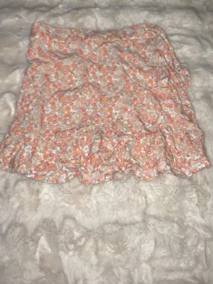 Blommig volangkjol i pastellfärger - Söt kortkjol med blommigt mönster i ljusa pastellfärger som rosa, vit och orange. Kjolen har volangdetalj längst ner och är gjord i ett lätt och luftigt material, perfekt för varmare dagar. Resår i midjan för skön passform. Storlek 146/152. Pris 20kr