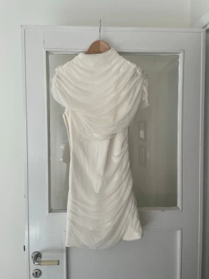 Vit omlott kortklänning - Säljer en vit kortklänning med omlott-detaljer och draperad effekt. Klänningen har hög krage och är ärmlös, vilket ger en elegant och modern look. Perfekt för dig som vill sticka ut med en stilren och trendig outfit.