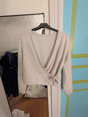 Beige omlottstickad tröja - Säljer en beige stickad omlottopp med långa ärmar och knytband i sidan. Tröjan har en djup V-ringning och ribbade muddar vid ärmslut och nederkant. Perfekt för lager på lager och enkel att styla till olika outfits. 