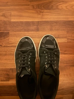Lanvin - Snygga svarta sneakers med coola lackdetaljer på tån och klassisk vit sula. Skorna har snörning med metallhål och ovandel i mockaliknande material. Perfekta för dig som gillar en stilren men ändå edgy look. Dem är riktiga och riktigt snygga🤩🤩