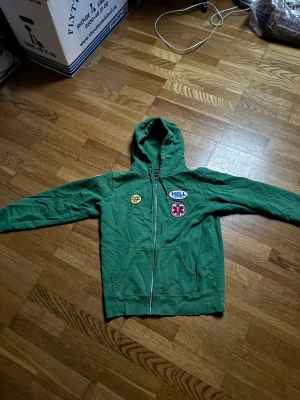 Supreme hell hysteric zip hoddie  - Grön hoodie med dragkedja framtill och flera patches, bland annat en med texten 'HELL', en rund gul patch och en stor patch med skelett-händer på ryggen. Amerikansk flagga på ena ärmen. Mjuk bomullskänsla och kängurufickor framtill.