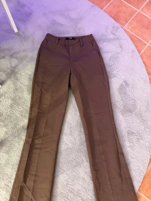 Bruna kostymbyxor från Bik Bok - Snygga bruna kostymbyxor från Bik Bok med raka ben och pressveck, lite utsvängning. Byxorna har hällor för skärp och stängs med knapp och dragkedja. Perfekta för en stilren och trendig look.