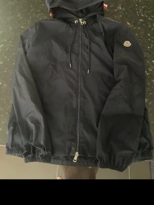 Blå  vindjacka med huva från Moncler - Snygg blå vindjacka från Moncler med dragkedja framtill, huva med dragsko och elastiska muddar. Jackan har Moncler-logga på ärmen och en diskret rödvit och blå rand på huvan. Perfekt för dig som vill ha en stilren och sportig look. Den passar M mer än L men funkar till båda !