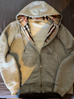 Grå hoodie från Burberry med dragkedja - Snygg grå hoodie från Burberry med dragkedja framtill och klassiskt rutigt foder i huvan. Hoodien har fickor på magen, ribbade muddar och justerbar dragsko i huvan. Perfekt för dig som gillar stilrena plagg med en twist av lyx.