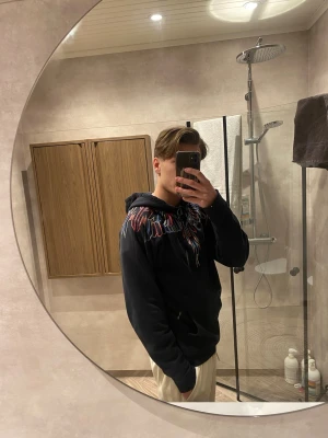 Svart Marcelo Burlon hoodie med färgglatt tryck - Säljer en svart hoodie från Marcelo Burlon med ett färgglatt grafiskt tryck runt halsen och över axlarna. Hoodien har huva med dragsko och en avslappnad passform. Perfekt för dig som vill sticka ut med en unik design.