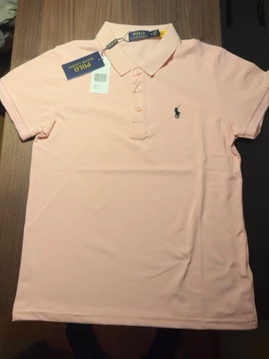 Ljusrosa pikétröja från Ralph Lauren - Säljer en ljusrosa pikétröja från Ralph Lauren med klassisk krage och tre knappar framtill. Tröjan har korta ärmar och den ikoniska broderade loggan på bröstet. Perfekt för en clean och stilren look.