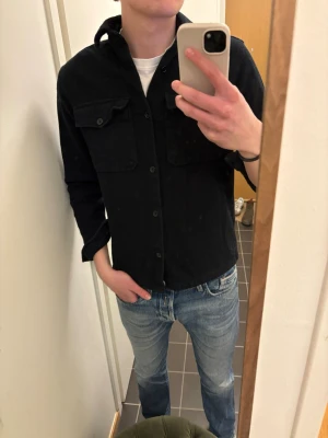 Svart overshirt från Jack & Jones - Stilren svart overshirt från Jack & Jones Premium med två stora bröstfickor och knäppning framtill. Skjortan har klassisk krage och är tillverkad i ett mjukt tyg som känns skönt mot huden. Perfekt att bära över en t-shirt för en avslappnad look.