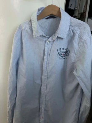 Blårandig skjorta från Guess - Säljer en blårandig skjorta från Guess med Los Angeles-brodyr på bröstet. Skjortan har klassisk krage, knäppning framtill och långa ärmar. Perfekt för dig som gillar en clean och stilren look.