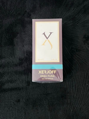 Xerjoff Erba Pura Eau de Parfum 100ml - Lyxig parfym från Xerjoff, Erba Pura Eau de Parfum i 100 ml. Förpackningen är elegant i lila med gulddetaljer och en turkos rand. Doften är känd för sin fräscha och fruktiga karaktär, och flaskan är tillverkad i glas.
