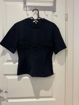 Svart topp med virkad detalj - Cool svart topp med korta ärmar och unik virkad detalj över bysten. T-shirten har rund halsringning och är gjord i mjukt bomullsmaterial. Passformen är normal och toppen har en stilren, modern look som sticker ut.