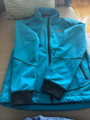 Blå träningsjacka från Swix - Snygg blå träningsjacka från Swix med hel dragkedja framtill och svarta detaljer vid muddar och fickor. Jackan har hög krage och två sidofickor med dragkedja. Perfekt för löpning eller utomhusträning. Materialet känns mjukt och funktionellt.