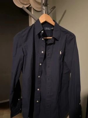 Mörkblå skjorta från Polo Ralph Lauren - Klassisk mörkblå skjorta från Polo Ralph Lauren med vit broderad logga på bröstet. Skjortan har lång ärm, knappar framtill och är tillverkad i bomull. Perfekt för en clean och stilren look.