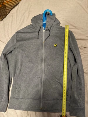 Grå zip hoodie från Lyle & Scott - Snygg grå hoodie från Lyle & Scott i storlek M. Dragkedja framtill, huva med snören och den klassiska gula loggan på bröstet. Tillverkad i mjukt material som känns skönt mot huden. Perfekt för en avslappnad och stilren look.