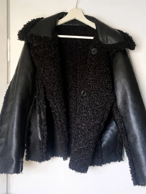 Vändbar jacka från H&M - svart, strl S/M - Snygg vändbar jacka från H&M i väldigt bra skick. Ena sidan är ytbehandlad, slät kvalitet och den andra är i mjuk pile/teddy - perfekt om man vill kunna variera stil. Jackan har krage, knäppning framtill och långa ärmar.   Storleken är oklar då lappen saknas. Jag brukar ha M i jackor och denna sitter något tajt på mig, så den passar troligtvis en S eller en mindre M.