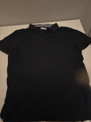 Mörkblå piké med krage - En klassisk svart pikétröja med krage och knappar framtill. Tröjan har korta ärmar och en enkel, stilren design som passar till det mesta. Perfekt för dig som gillar en clean och tidlös look.