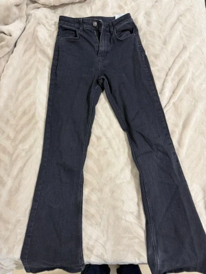 Svarta bootcut jeans med slitning - Säljer ett par svarta bootcut jeans med hög midja och klassisk femficksdesign. Jeansen har en tydlig slitning och trasig detalj vid ena benslutet för en edgy look. Materialet är denim med lätt stretch för skön passform.