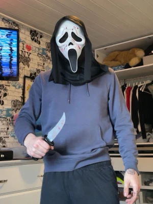 Scream maskeradmask och fejkkniv - Säljer en klassisk Scream-mask i vit plast med svart tyg och blodstänk, samt en fejkad kniv i plast med blodiga detaljer. Perfekt för halloween eller cosplay. Masken täcker hela ansiktet och har svart huva.