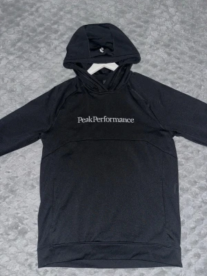 Svart hoodie från Peak Performance - Säljer en svart hoodie från Peak Performance med stor vit logga och text 'Est. 1986' på bröstet. Klassisk känguruficka framtill och justerbar huva med snören. 