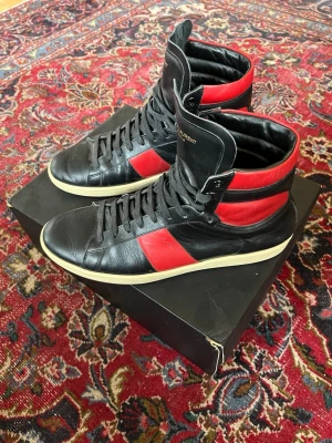 Saint Laurent svarta och röda sneakers - Snygga high-top sneakers från Saint Laurent i modellen sl/02h. Storlek 44 och har riktigt bra skick. Boxen ingår ej! Om du har någon fundering är det bara att fråga 